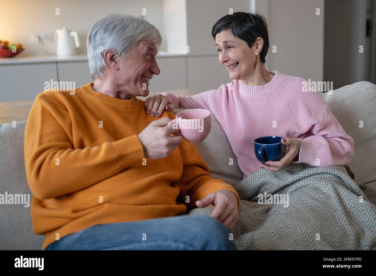 Fröhliches Seniorenehepaar teilt Tee und Unterhaltung, entspannt zusammen auf dem Sofa zu Hause drinnen Stockfoto