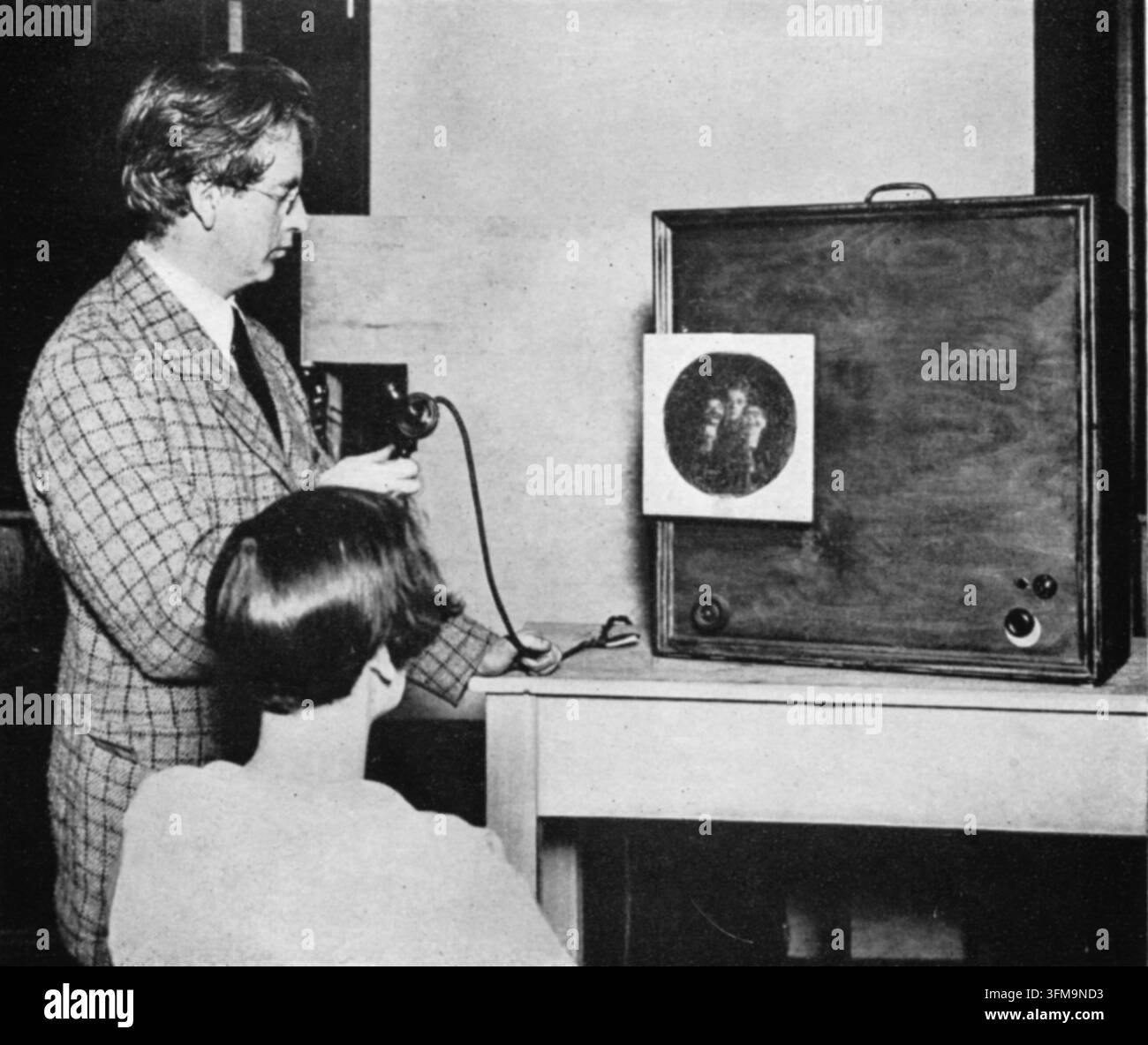 Ein Foto von John Logie Baird (1888–1946), der neben einem frühen Fernsehsender steht. Der schottische Elektroingenieur und Fernsehpionier ist während eines Experiments mit seinem mechanischen Fernsehsystem zu sehen, das oft als „drahtlose Sicht“ bezeichnet wird und eine sich drehende Nipkow-Schibe zur Abtastung und Übertragung von Bildern verwendete. Der Apparat zeigt einen großen Holzschrank mit einer kleinen runden Öffnung, die ein übertragenes Bild darstellt, während ein Beobachter im Vordergrund die Live-Übertragung verfolgt. Ca. 1926. Stockfoto