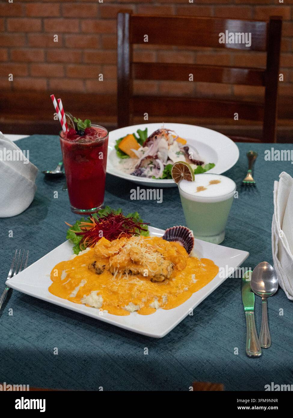 Gericht auf weißem Teller mit orangefarbener Sauce. Frischer Salat neben einem lebendigen roten Getränk. Tisch für zwei Personen mit sorgfältig angeordnetem Besteck. Grüne Tischdecke a Stockfoto