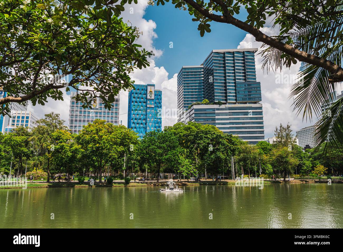 Bangkok, Thailand, 3. Mai 2026: Der üppige Lumphini-Park umrahmt einen ruhigen See, ein kleines Boot und die moderne Skyline von Bangkok unter einem strahlend blauen Himmel. Stockfoto