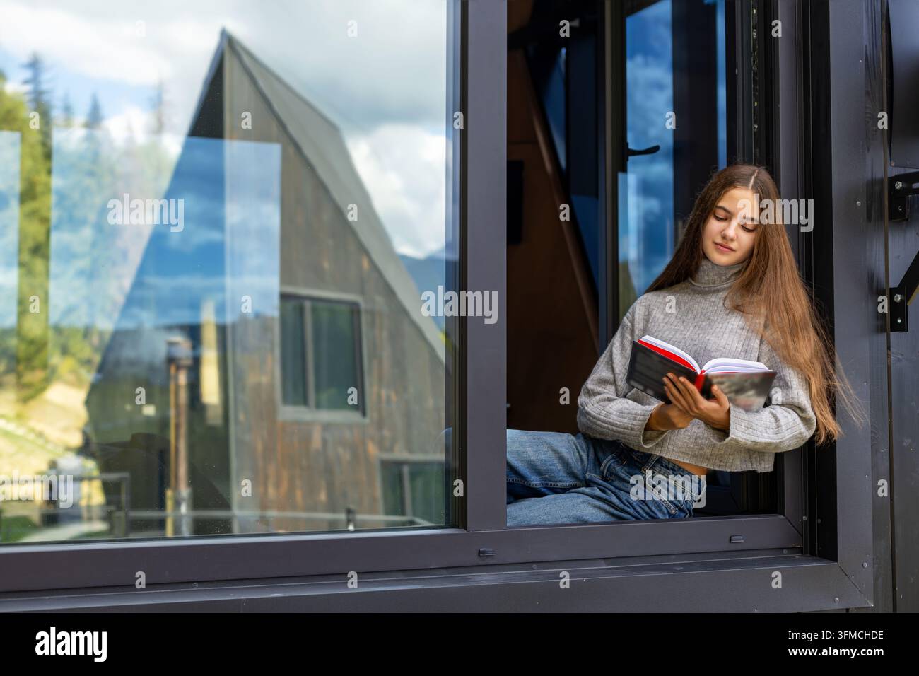 Junge Frau liest ein Buch an einem offenen Fenster eines modernen Hauses, friedlicher Freizeitmoment mit Berglandschaft und Reflexionen in großem Glas. Stockfoto