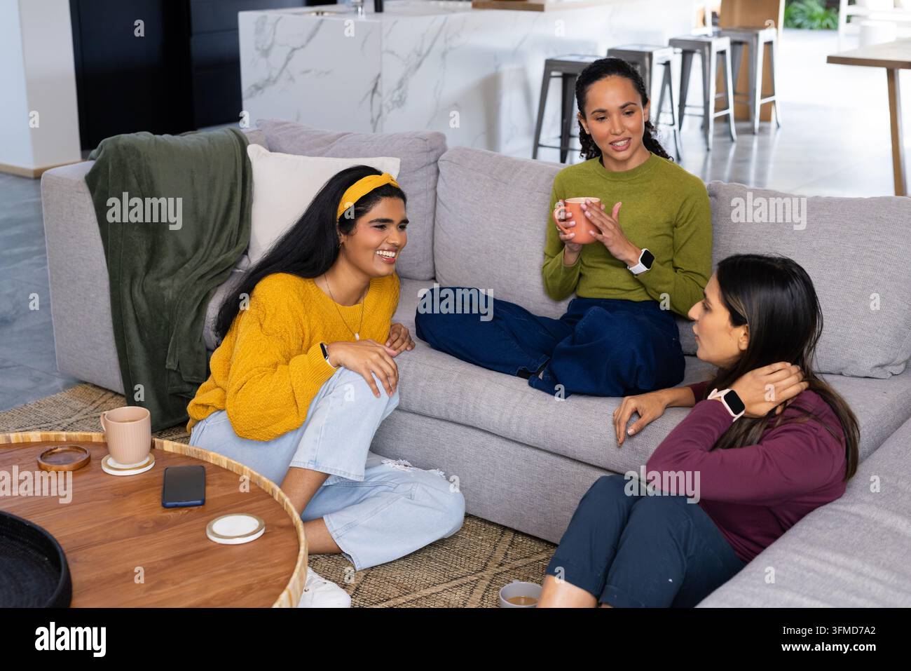 Verschiedene Freundinnen, die sich auf einem grauen Sofa zu Hause unterhalten und aus Tassen trinken, mit einem Telefon auf dem Tisch Stockfoto