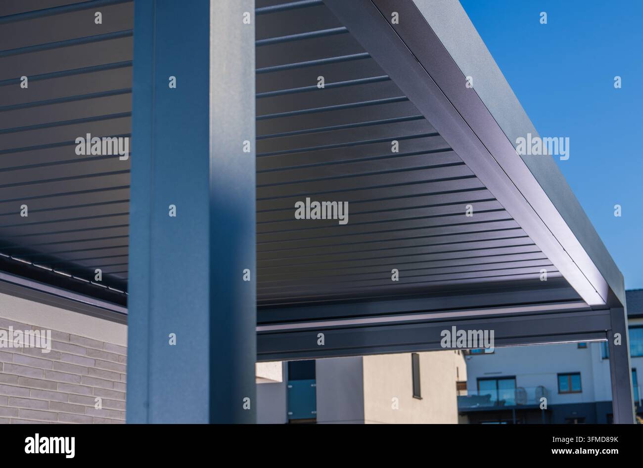 Architektonisches Detail eines dunklen Metallpergolenrahmens und eines Lamellenhimmels neben einem modernen Haus unter einem klaren blauen Himmel. Stockfoto