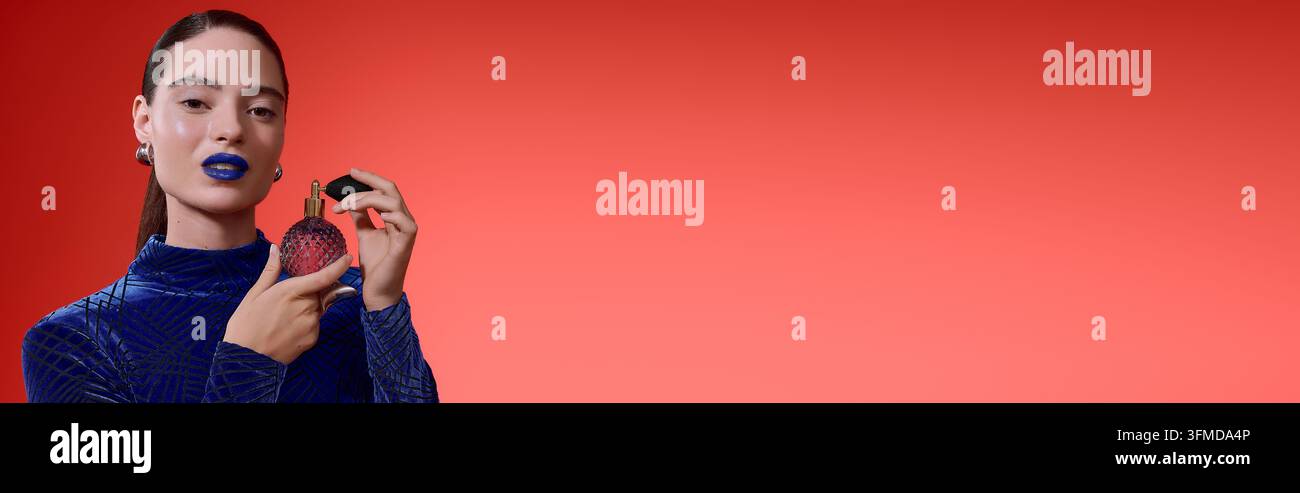 Eine junge Frau präsentiert auffälligen blauen Lippenstift und einen samtenen Rollkragen-Bodysuit, während sie eine Parfümflasche und ein Banner hält Stockfoto