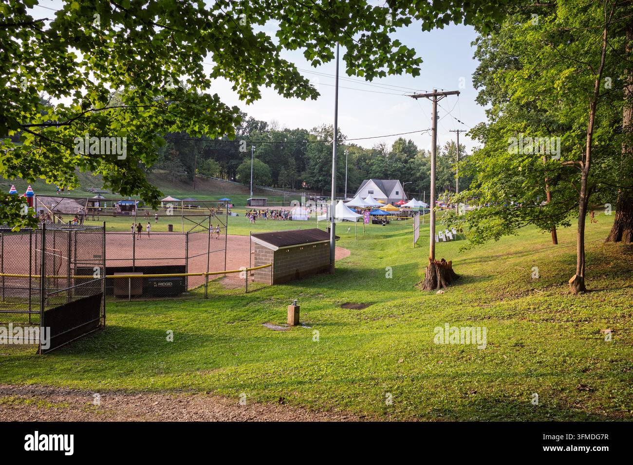 Pleasant Hills Pennsylvania USA-08 08 2026: Ein leerer Baseball-Backstop und ein grasbewachsenen Hügel umrahmen ein fernes Sommerfest im Freien mit weiß Stockfoto
