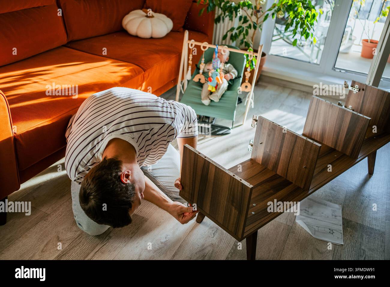 Vater baut neues TV-Ständer-Möbel auf dem Boden, folgt sorgfältig den Anweisungen, während sein Baby von einem Wippgestell im hell erleuchteten Wohnzimmer aus zusieht und ein Familienheim erschafft Stockfoto