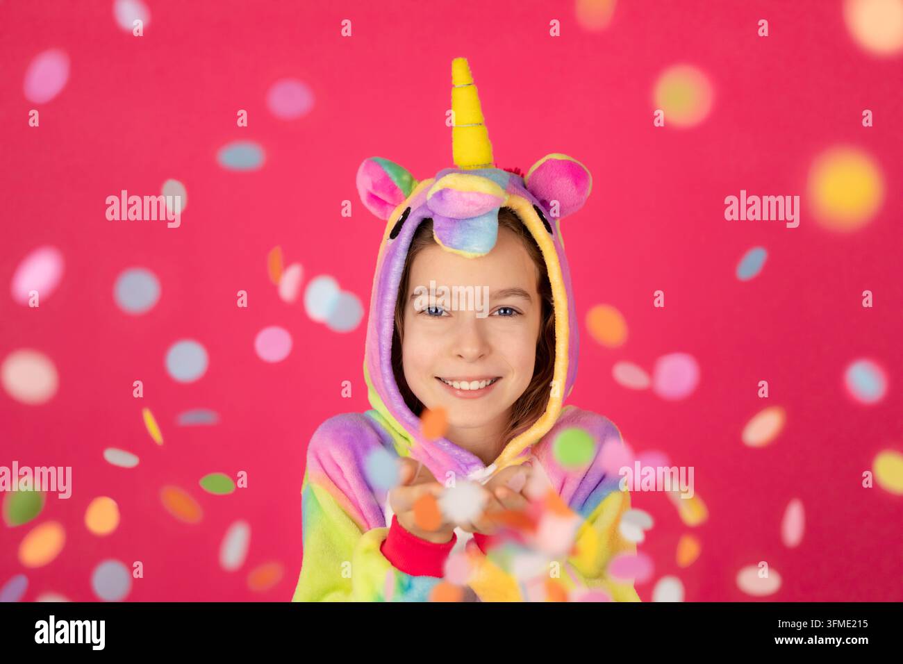 Schickes Mädchen in Einhornkostüm, das Konfetti bläst. Porträt eines lustigen Kindes vor rosa Hintergrund. Weihnachtliches Urlaubskonzept Stockfoto
