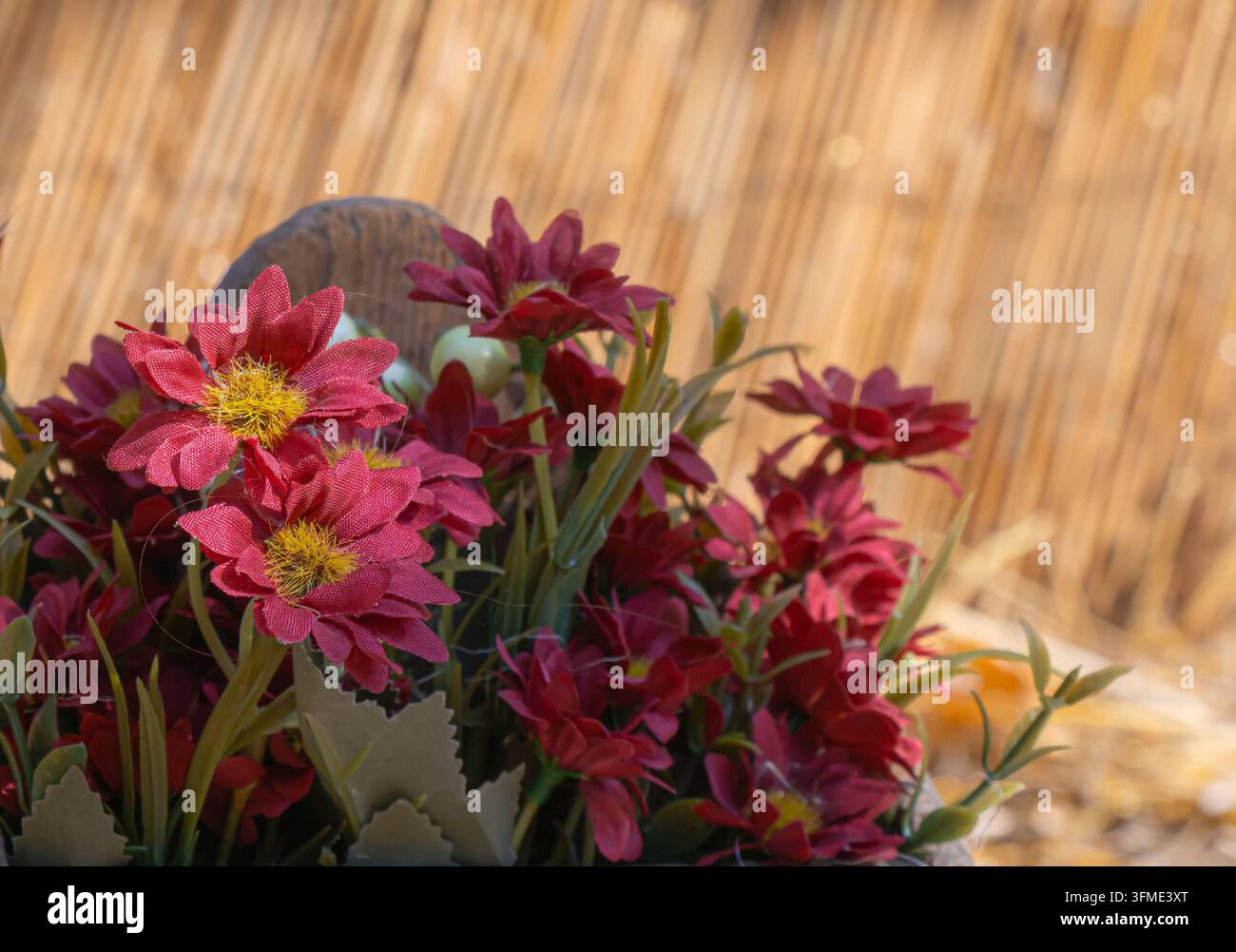 Nahaufnahme von roten künstlichen Stoffblumen mit gelben Mittelpunkten und grünen Blättern, angeordnet vor warmem Stroh für rustikales Deko- und Basteldesign. Stockfoto