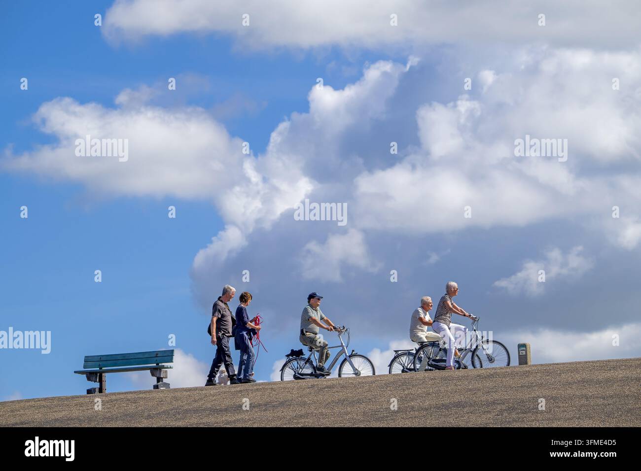 Rentner fahren auf Fahrrädern / Rädern an einem älteren Paar vorbei, das seinen Hund über den Deich führt an einem bewölkten Tag im Spätsommer / Frühherbst Stockfoto