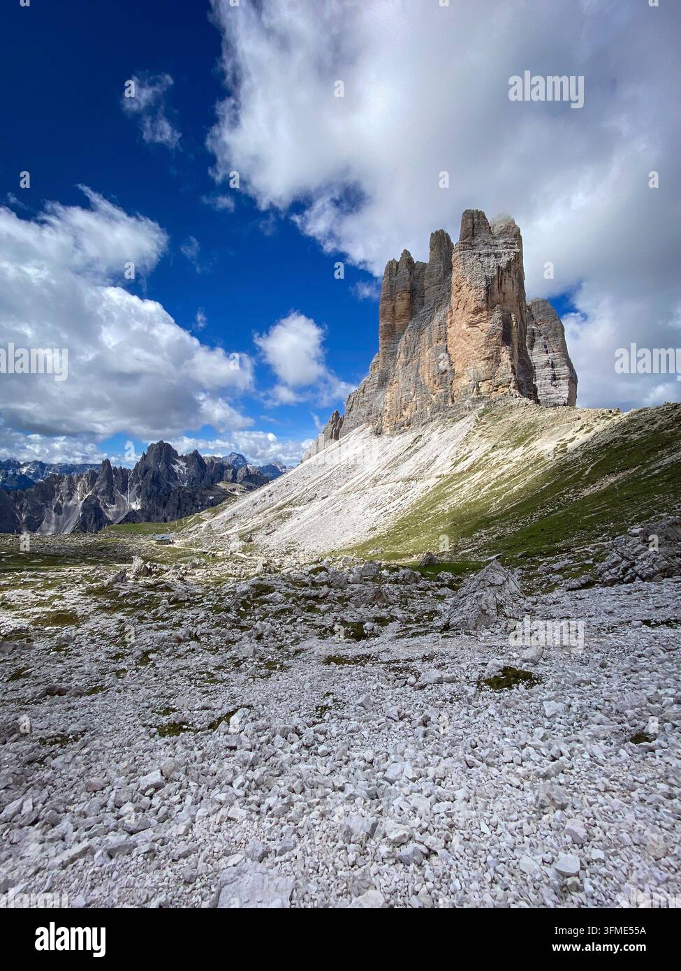 Drei Zinnen von Lavaredo mit der Cima Grande rechts und den fernen Cadini di Misurina, heller Kalkstein-Schutt und grüne Hänge vor blauem Himmel Stockfoto