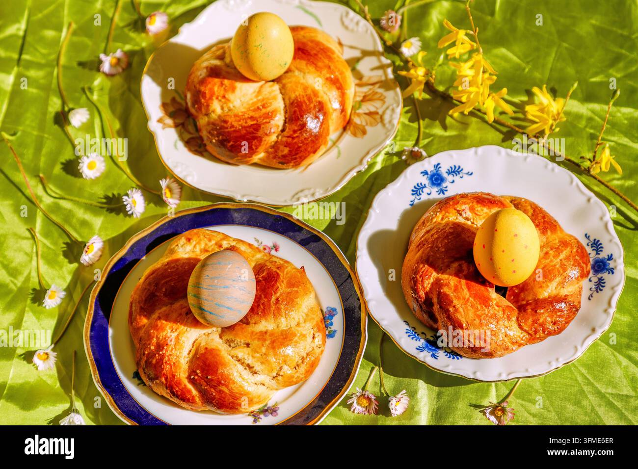 Drei Osterkränze mit farbigen Ostereiern auf bunten Tellern auf grüner Krapfen-Tischdecke mit Gänseblümchen und Forsythienzweig Stockfoto