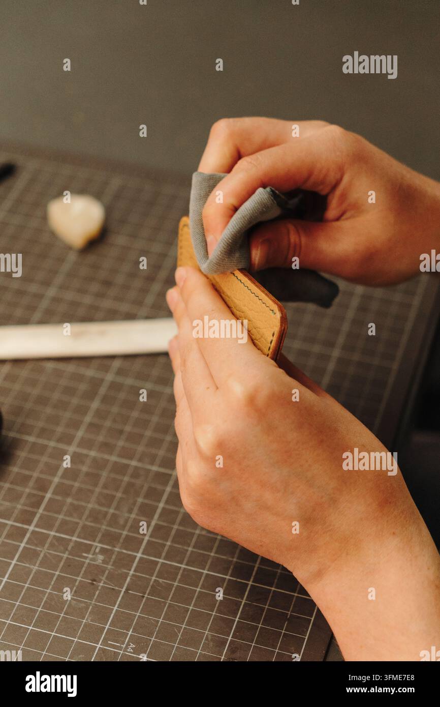 Hände polieren ein handgefertigtes Lederportemonnaie mit einem weichen Tuch. Der Abschlussprozess unterstreicht Produktpflege, Qualität und traditionelles Handwerk Stockfoto