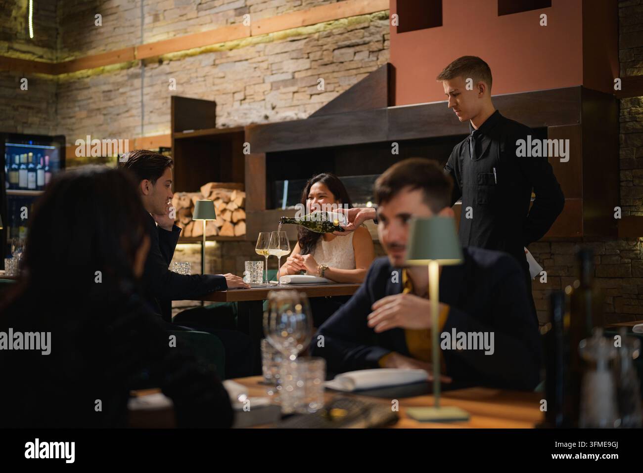 Kellner gießt Wein für ein Paar, das während eines romantischen Jahrestagsdinners in einem gemütlichen Luxusrestaurant anstößt, Seitenansicht. Stockfoto