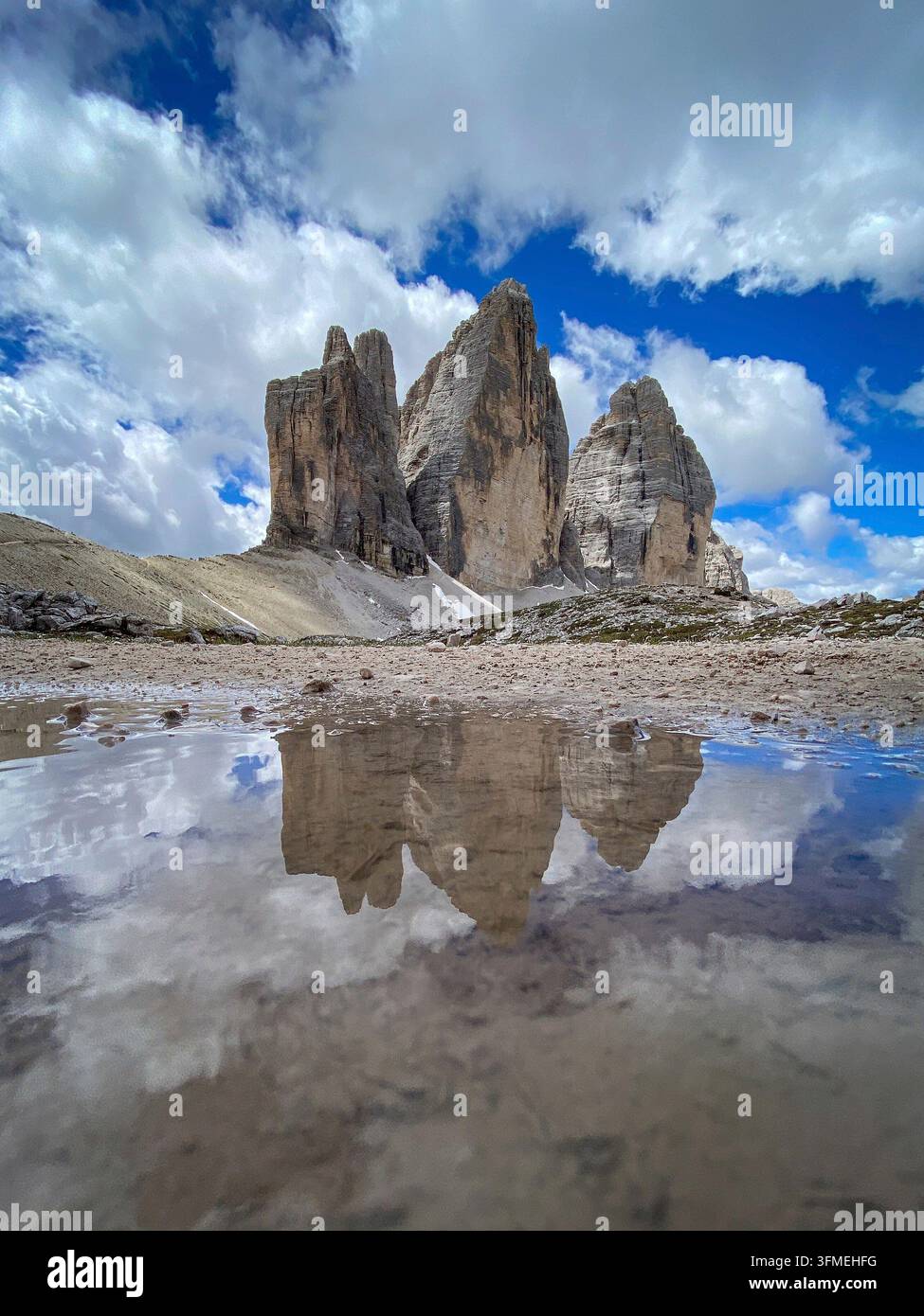 Niedrigwinklige Ansicht der Tre Cime di Lavaredo, die sich in einer Pfütze spiegeln, Cima Grande, Cima Ovest und Cima Piccola über hellem Kalkstein-Geröll vor blauem Himmel mit Stockfoto