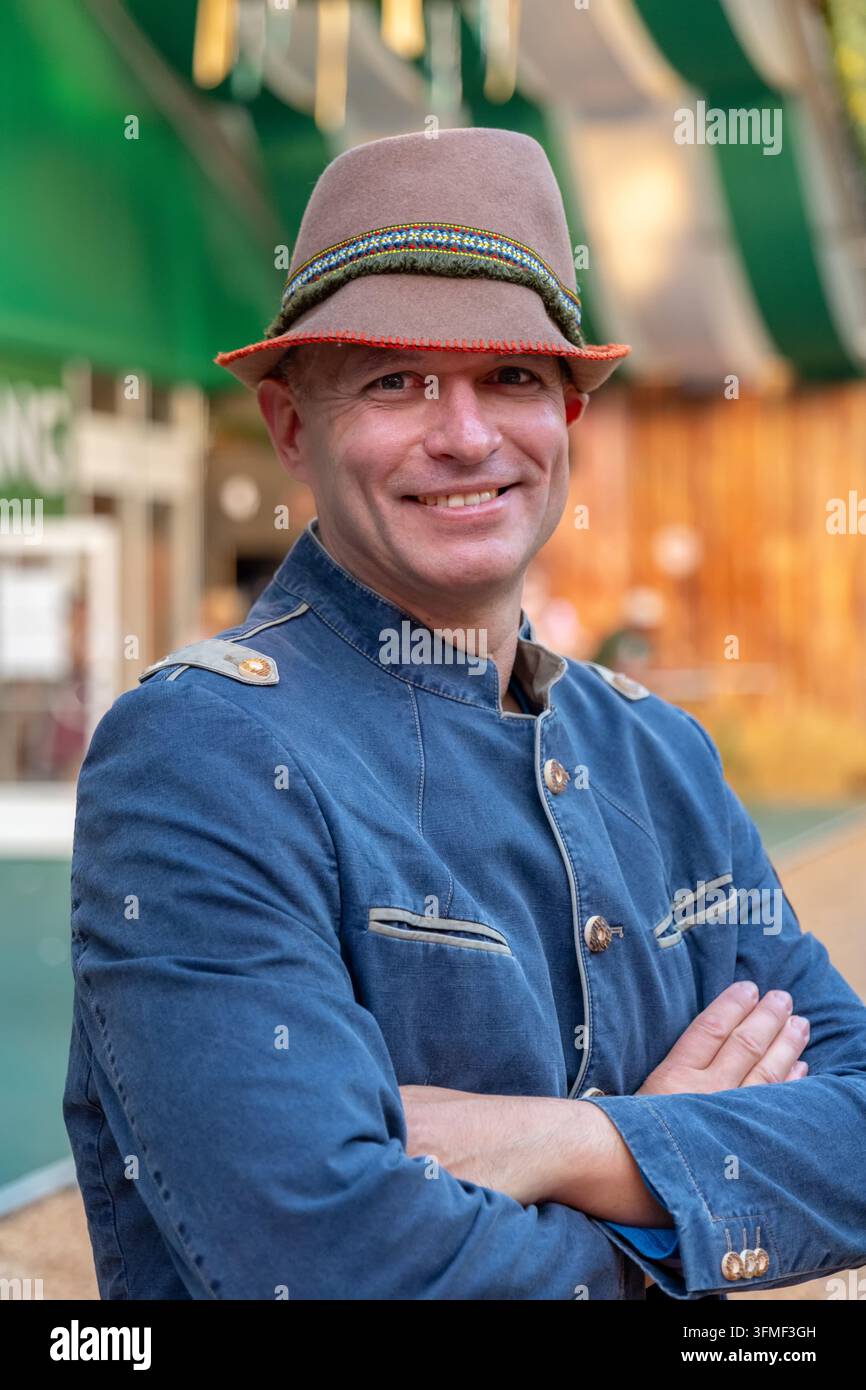 Ein lächelnder Mann in einem traditionellen Oktoberfest-Hut und blauer Jacke steht mit verschränkten Armen. Stockfoto