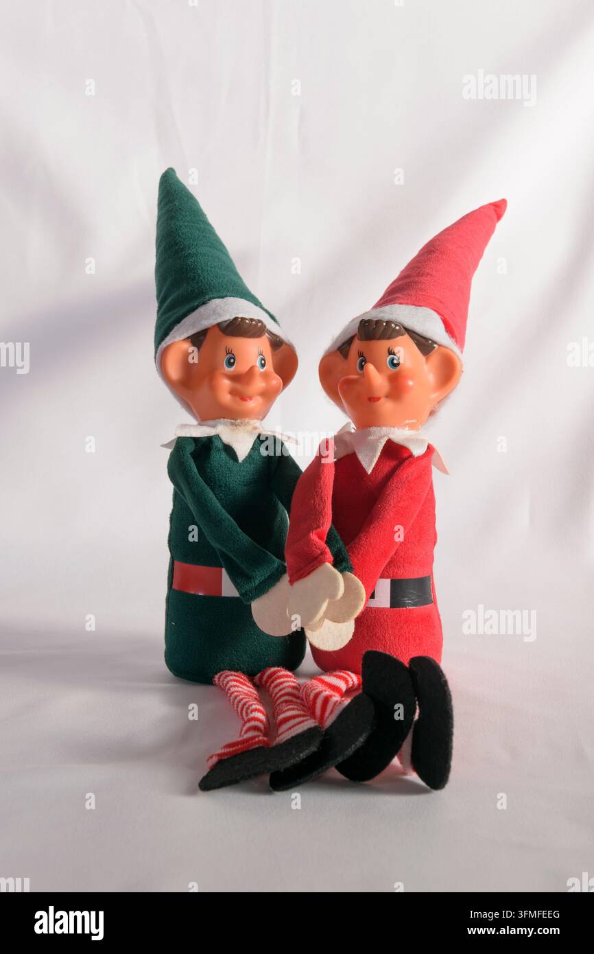 Studioaufnahme von zwei weihnachtlichen Elfenpüppchen im Vintage-Stil, eines grün und eines rot, die zusammen sitzen. Die Figuren haben lächelnde Plastikköpfe, spitze Stockfoto