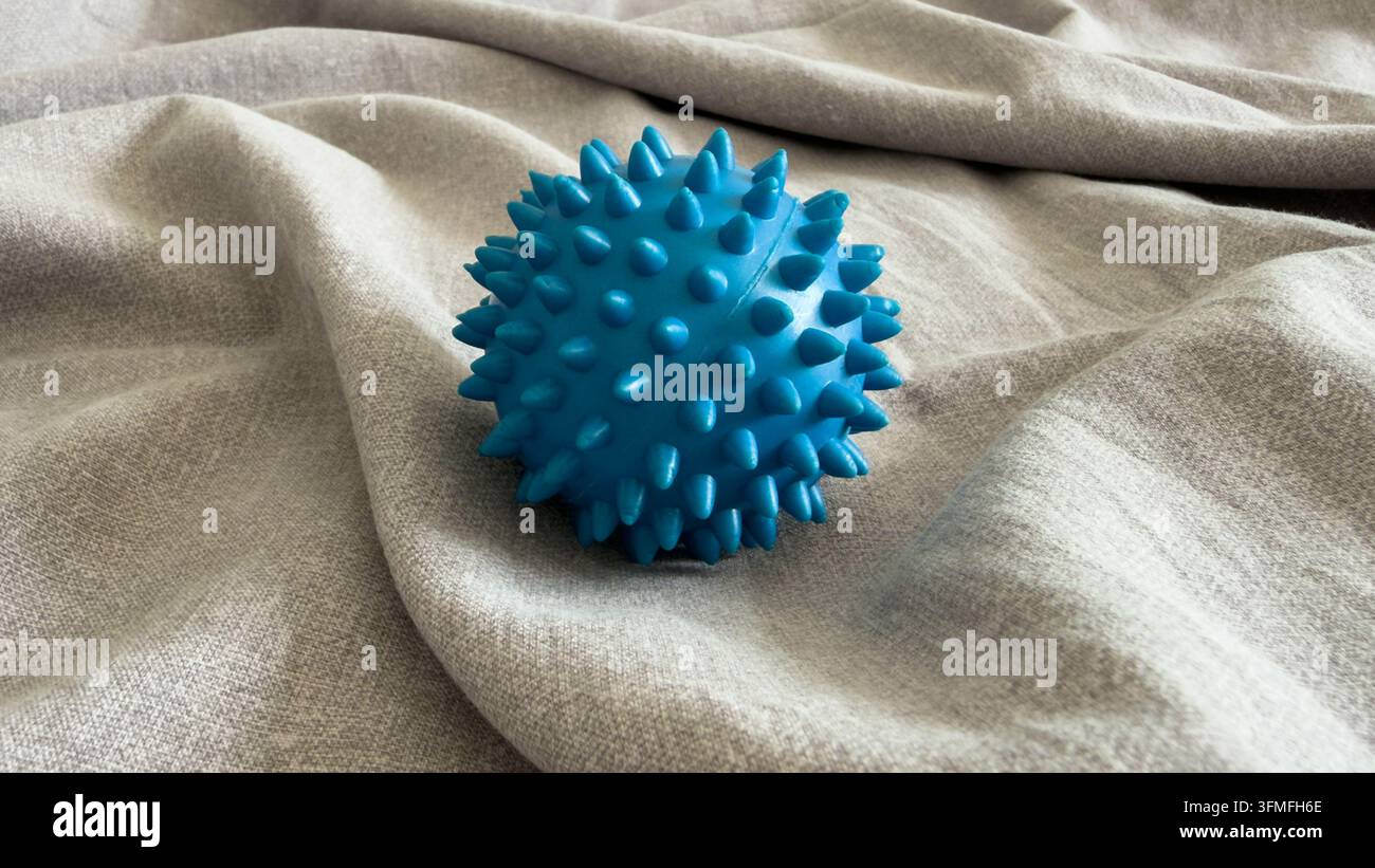 Nahaufnahme eines blauen stacheligen Akupressur-Massageballs auf strukturiertem grauem Stoff. Sensorisches stacheliges Ballwerkzeug für Myofaszialrelease, Triggerpunkttherapie, Physi Stockfoto