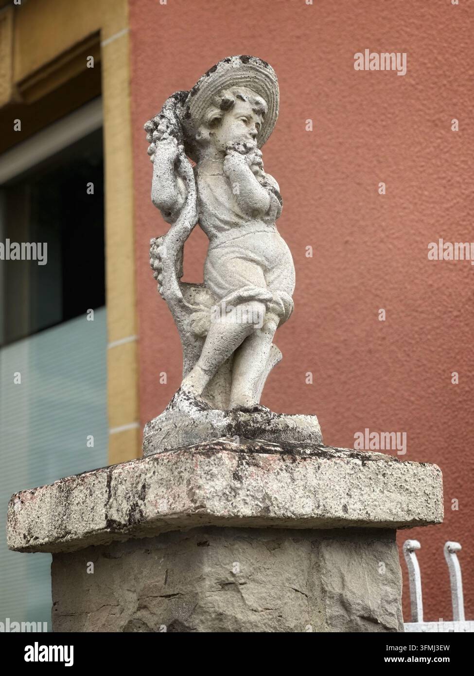 Alte Steinstatue einer Kinderfigur auf einem Pfeiler vor einer roten Wand. Verwitterte antike Steinskulptur eines Kindes mit Hut auf einem rustikalen Steinpfosten - Smartphone-aufgenommenes Stockfoto