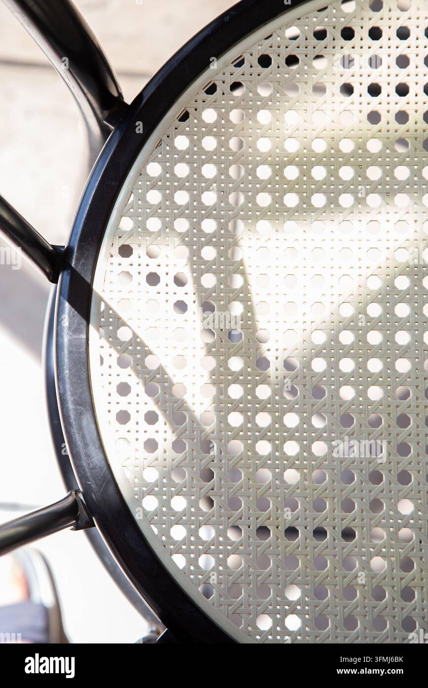 Nahaufnahme eines geflochtenen Korbstuhls mit schwarzem Metallrahmen, geometrischer Gittertextur, sonnenbeschienenen Innenschatten und neutralem modernem rustikalem Innenraum-Sitzdetail Stockfoto
