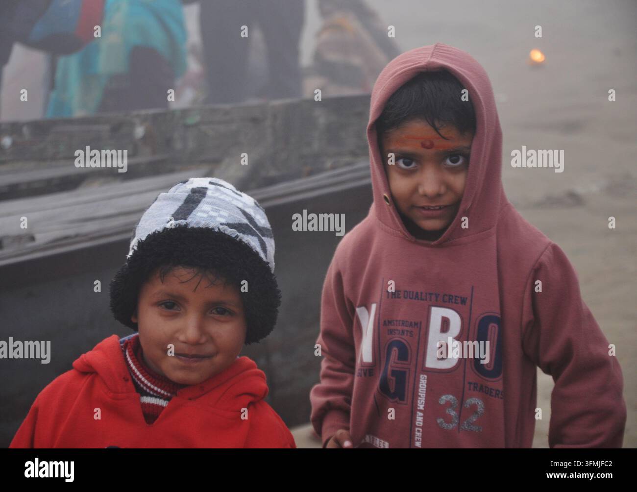 VARANASI, INDIEN - 23. JANUAR 2026: Zwei Kinder in Winterkleidung blicken neben einem Holzboot am Assi Ghat in dichtem Morgennebel in die Kamera Stockfoto
