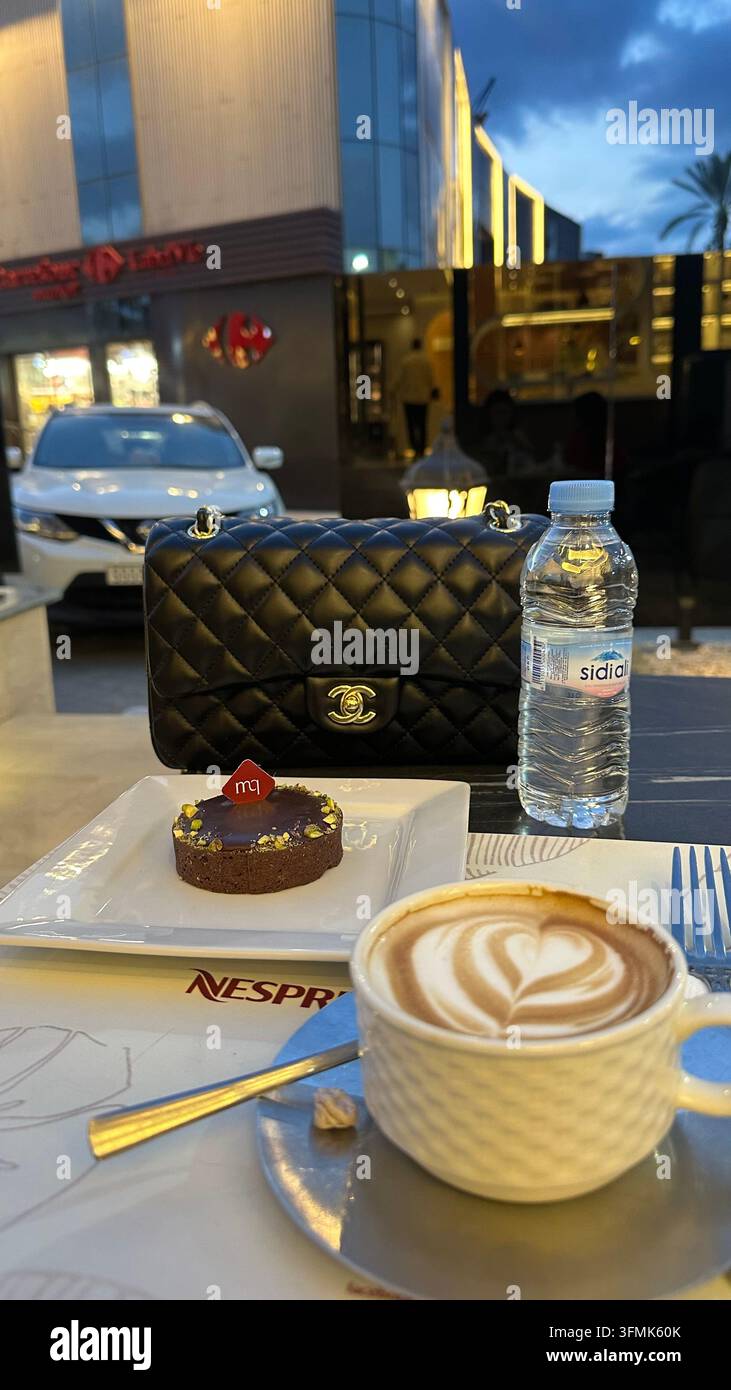 Tasse Kaffee mit herzförmiger Latte-Art und Schokoladendessert, serviert in einem Außencafé am Abend, mit Stadtlichtern im Hintergrund. Stockfoto