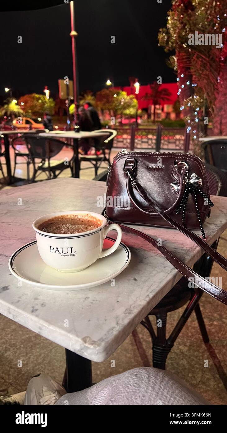 Tasse Kaffee und elegante burgunderrote Handtasche auf einem Marmortisch in einem Café im Freien bei Nacht, mit beleuchteten Palmen und einer lebhaften städtischen Umgebung in th - Smartphone-aufgenommenes Stockfoto