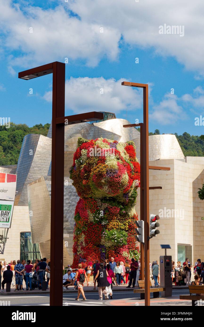 Jeff Koons' Puppy-Skulptur vor dem Guggenheim-Museum in Bilbao, eingerahmt zwischen zwei Laternenpfählen mit dem Museum im Hintergrund und Menschen, die die Straße überqueren. Stockfoto