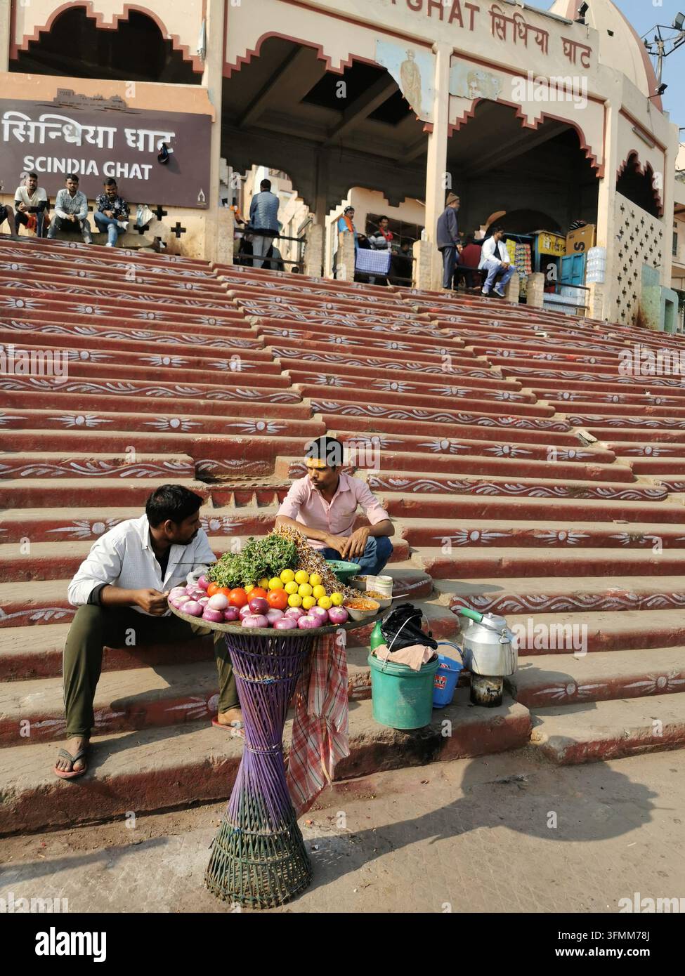 VARANASI, INDIEN - 22. JANUAR 2026: Zwei Männer sitzen auf rot gestrichenen Steinstufen, die mit weißen Mustern verziert sind, neben einem Stand, der Zwiebeln und Zitronen ausstellt Stockfoto