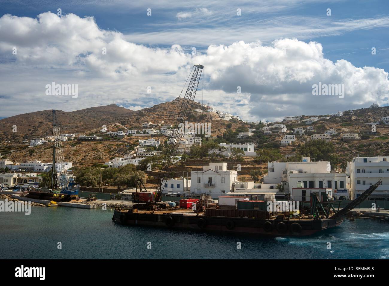 Der Hafen der Insel Ios, Griechenland, mit einem arbeitenden Kranschiff im Vordergrund, mit weißen kykladischen Häusern, die die Hügel unter dramatischen Wolken erklimmen Stockfoto