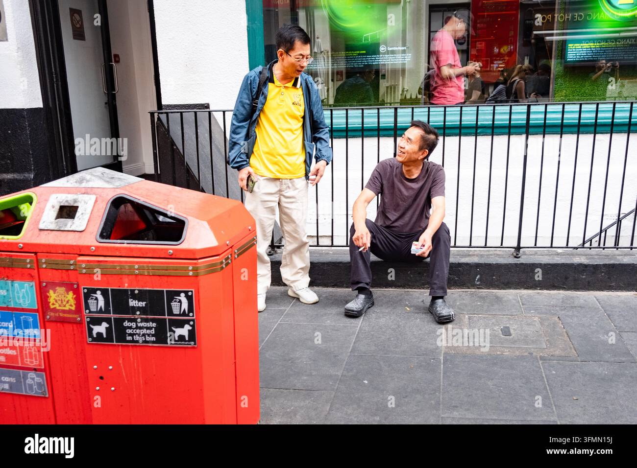 ZWEI MÄNNER MACHEN EINE PAUSE AUF EINEM STADTSTEIGER IN LONDON CHINATOWN, REISEBERICHT, LONDON, ENGLAND: Ein Mann sitzt auf dem Boden und raucht eine Zigarette, während ein anderer Mann neben ihm auf einem Stadtsteig neben einem roten Mülleimer steht und mit ihm spricht. INFO: Londons Chinatown, eingebettet im lebendigen Herzen von Soho, ist ein geschäftiger kultureller Mittelpunkt, der für seine authentischen asiatischen Restaurants, traditionellen Bäckereien und neonbeleuchteten Straßen bekannt ist. Die ikonischen roten Laternen bieten eine perfekte Kulisse für Feinschmecker. Stockfoto