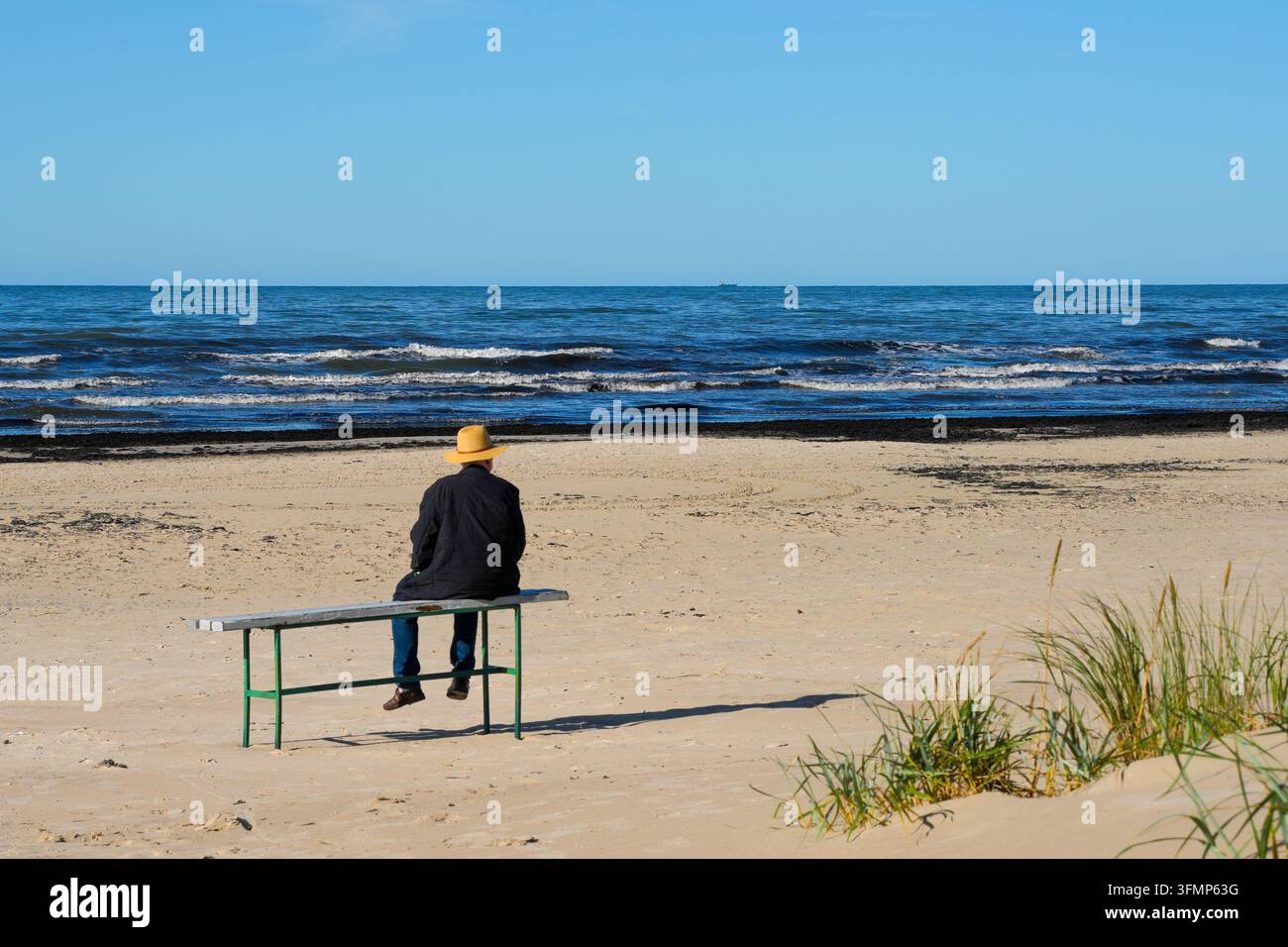Älterer Mann sitzt allein auf einer Bank und blickt auf das Meer von einem ruhigen Sandstrand im Herbst Stockfoto