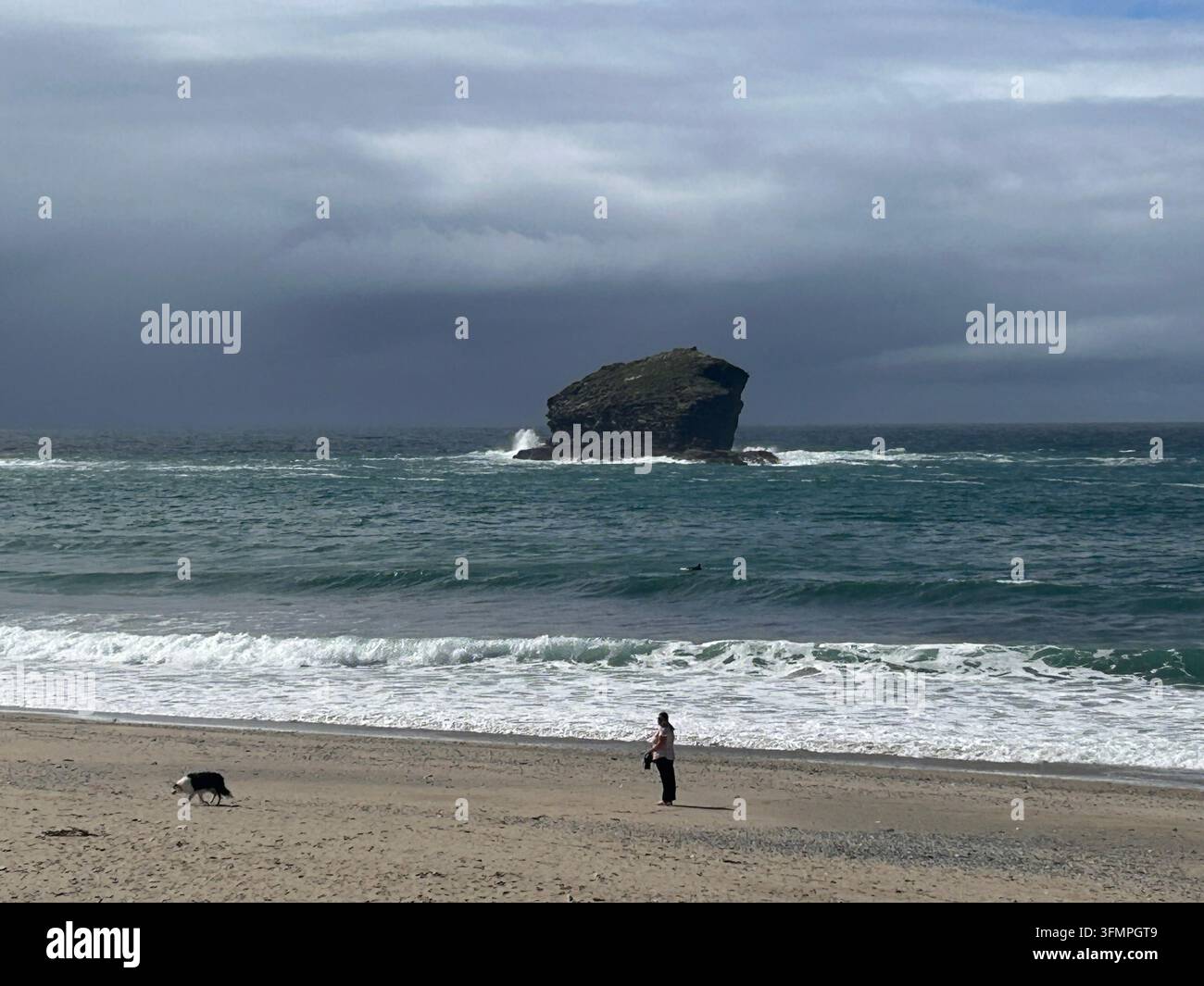 Wellen brechen um einen Offshore-Felsen am Strand von Portreath, mit einer Person und einem Hund auf dem Sand, Cornwall, England, Vereinigtes Königreich - Smartphone-aufgenommenes Stockfoto