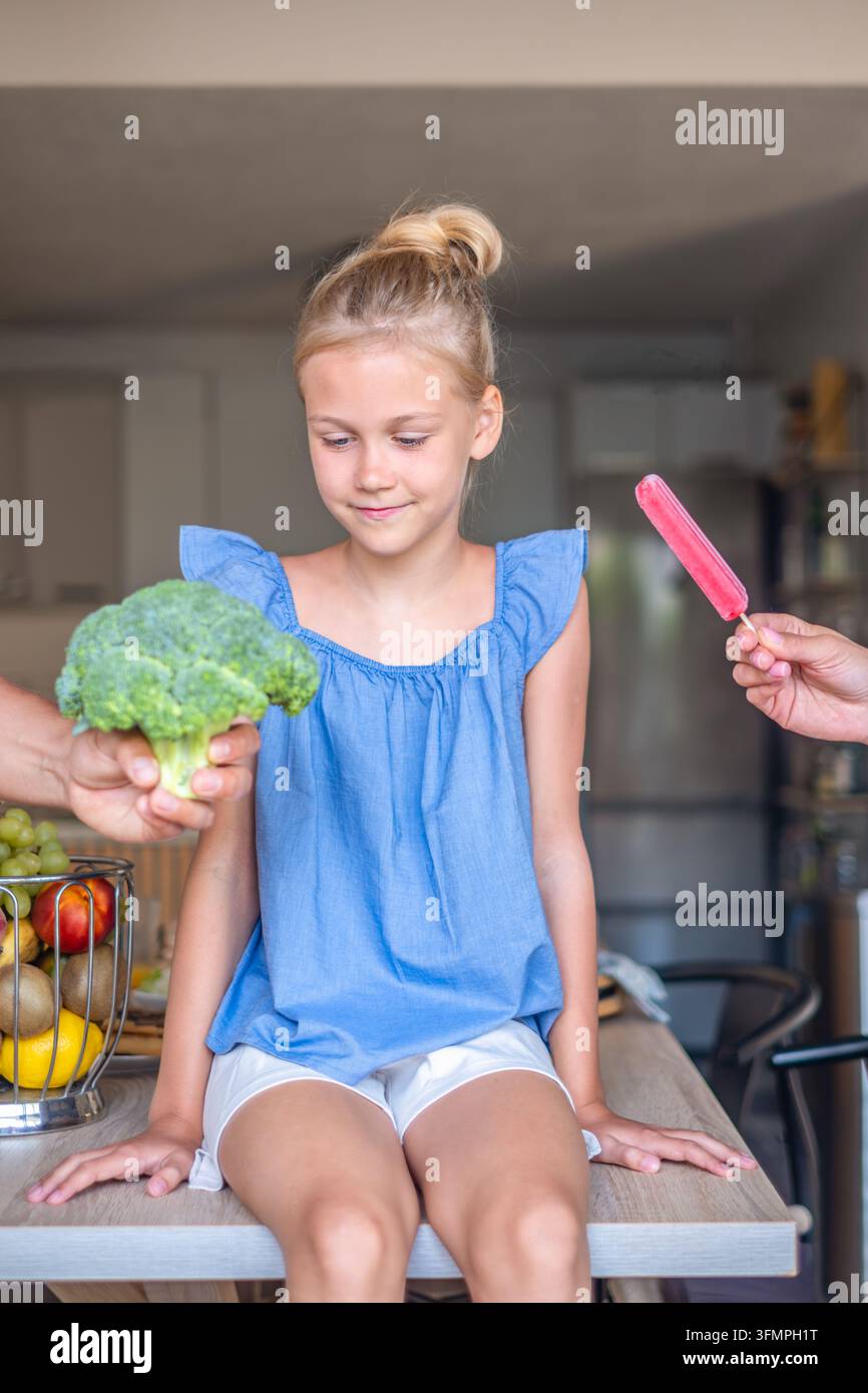 Blondes Mädchen wählt zwischen Brokkoli und Eis zu Hause . Hochwertiges Foto Stockfoto