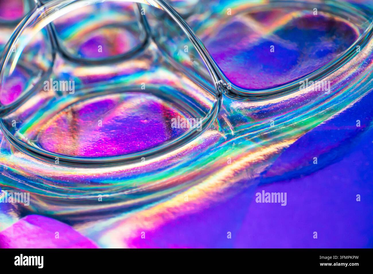 Makroabstrakt überlappender Glaslinsen, die lebendige irisierende Regenbogenreflexionen über eine leuchtende blaue, violette, rosa und türkisfarbene Oberfläche erzeugen Stockfoto