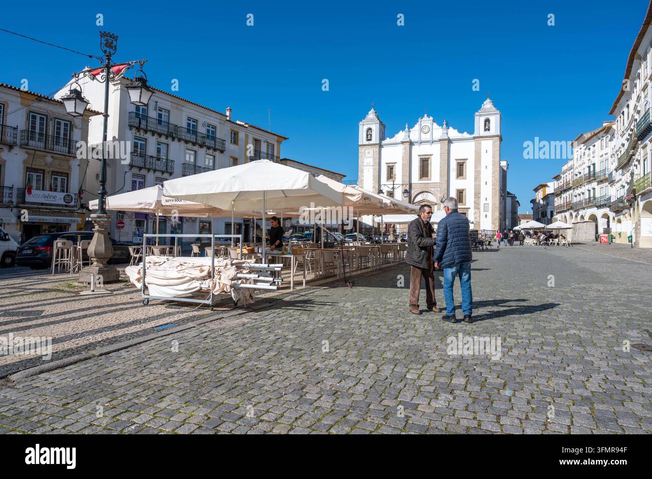 Évora, Portugal, 11-19-2024: Zwei Männer begrüßen sich auf Kopfsteinpflaster im historischen Stadtplatz mit Tischen und Stühlen, schattigem Restaurant im Freien Stockfoto