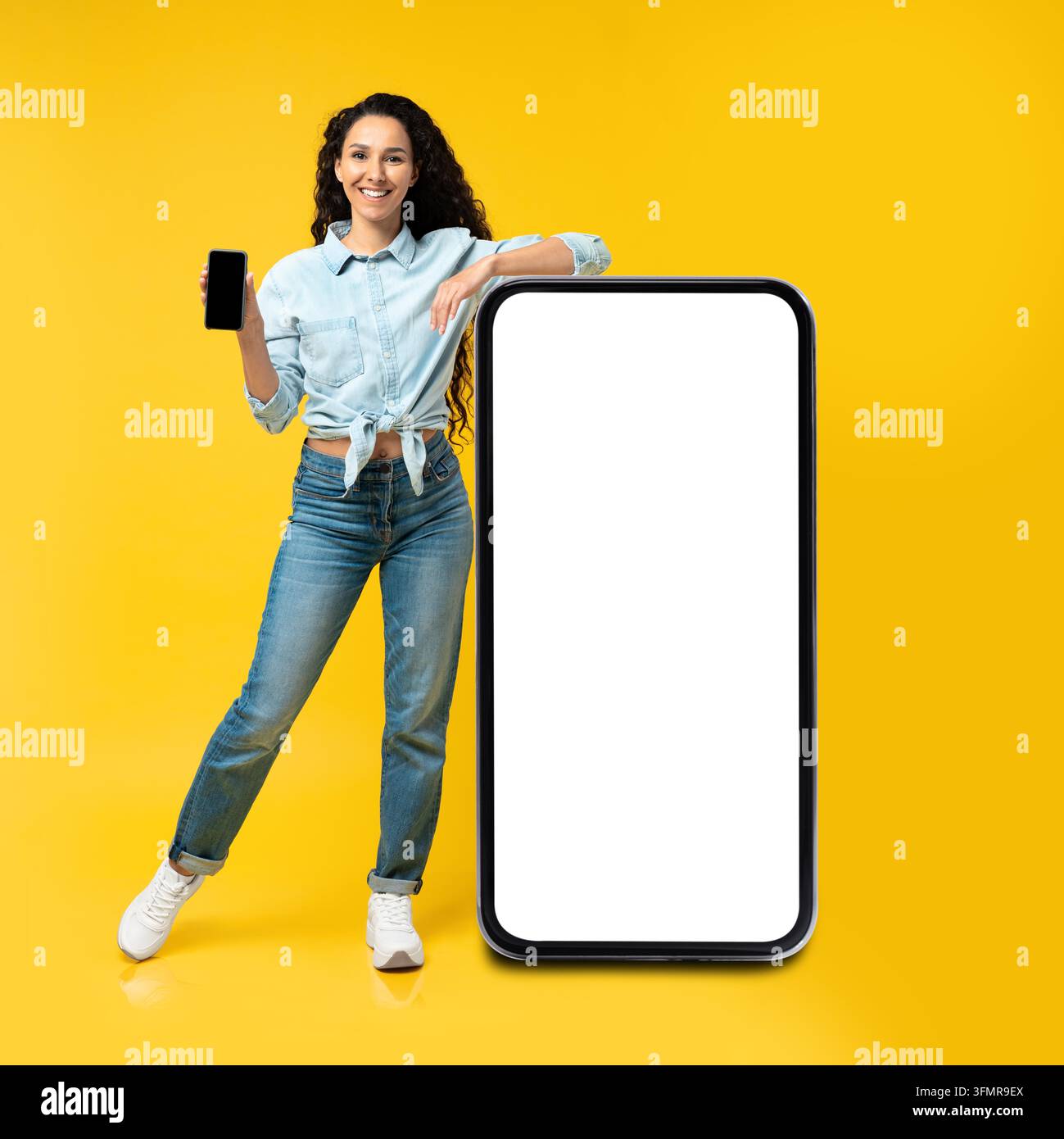 Lächelnde junge Frau hält Smartphone und posiert neben einem großen Mockup-Bildschirm, Studio-Setup Stockfoto