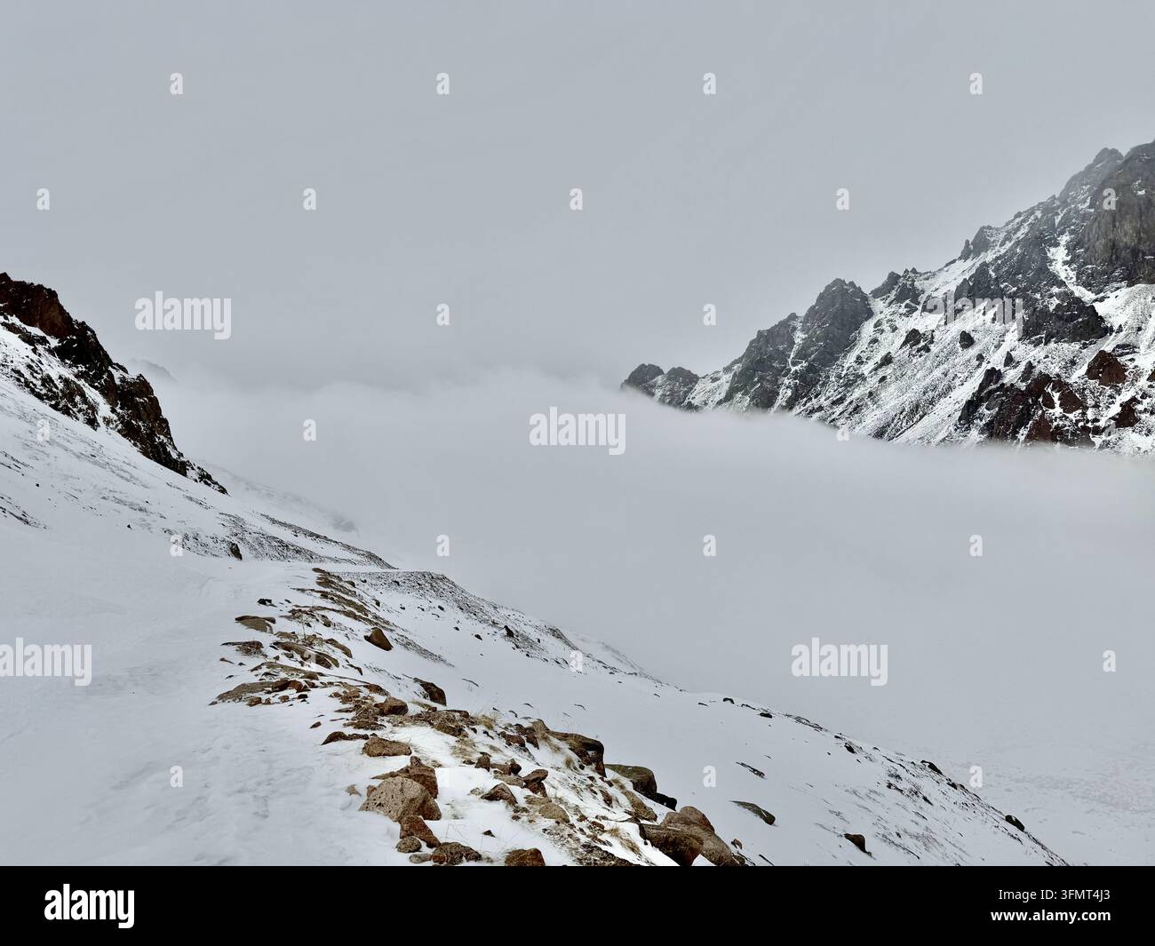 Dichter Nebel füllt ein schneebedecktes Bergtal unter schroffen Gipfeln nahe dem Tuyuksu-Gletscher im Ile Alatau, Kasachstan. - Smartphone-aufgenommenes Stockfoto