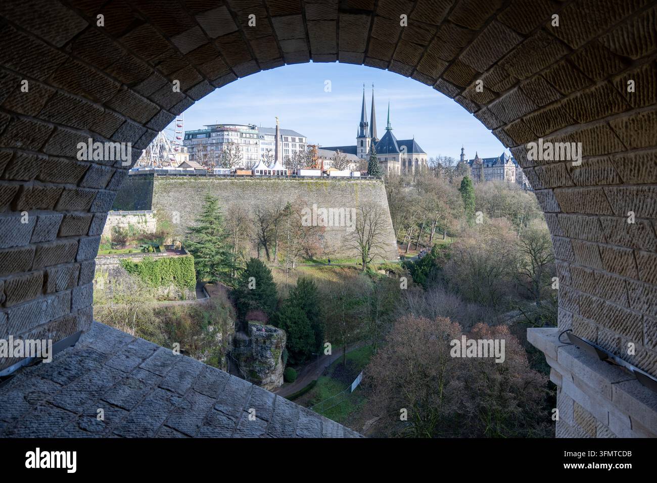 Ein Steinbogen umrahmt die Kathedrale der Stadt Luxemburg, historische Befestigungen und eine Winterlandschaft, wobei ein Riesenrad links sichtbar ist. Stockfoto