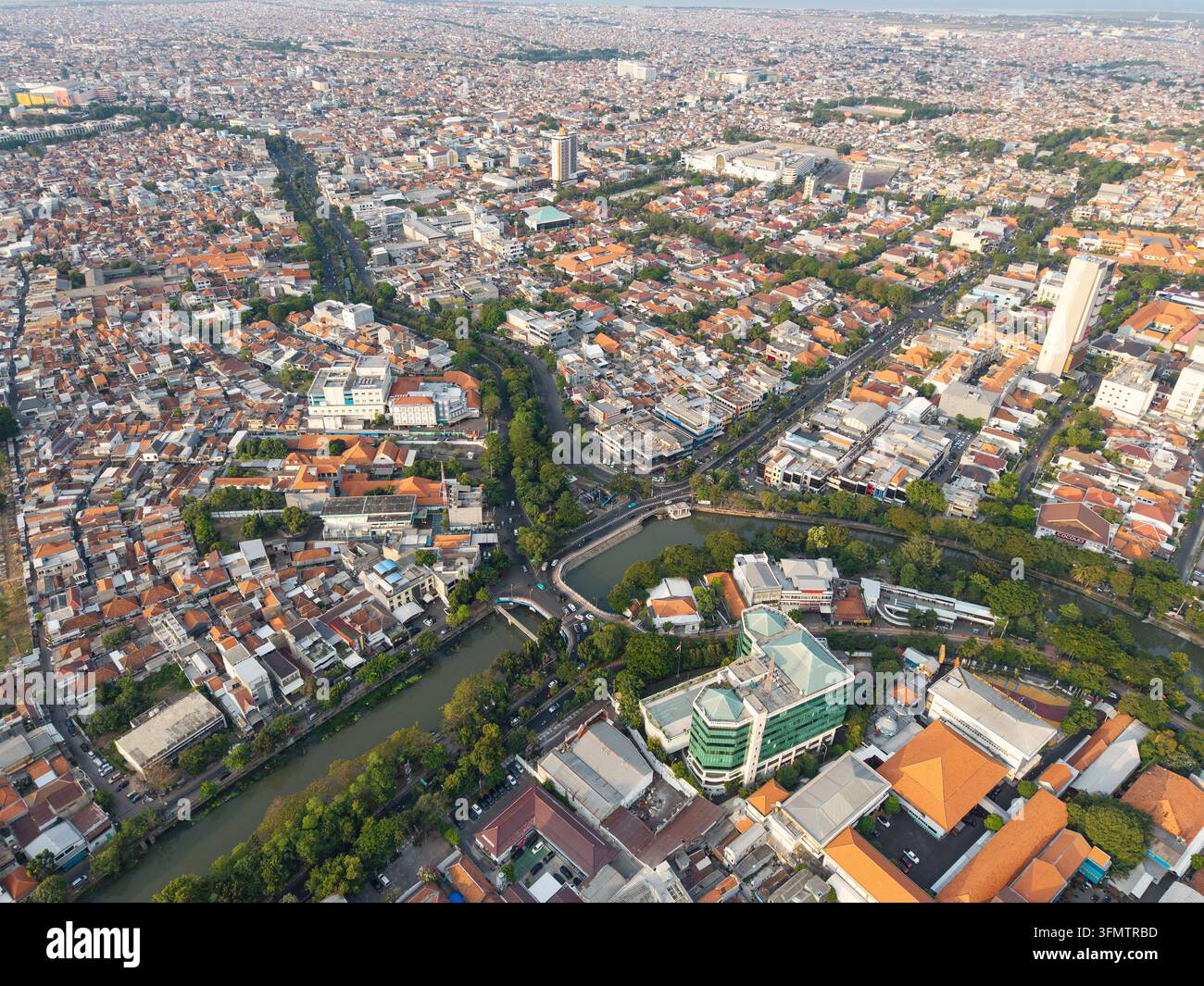 Luftpanorama am Tag über dichte Stadtviertel von Surabaya, Ziegeldächer, belebte Straßen, Gewerbegebäude, Grünflächen und ein städtisches Wasserlauf. Stockfoto