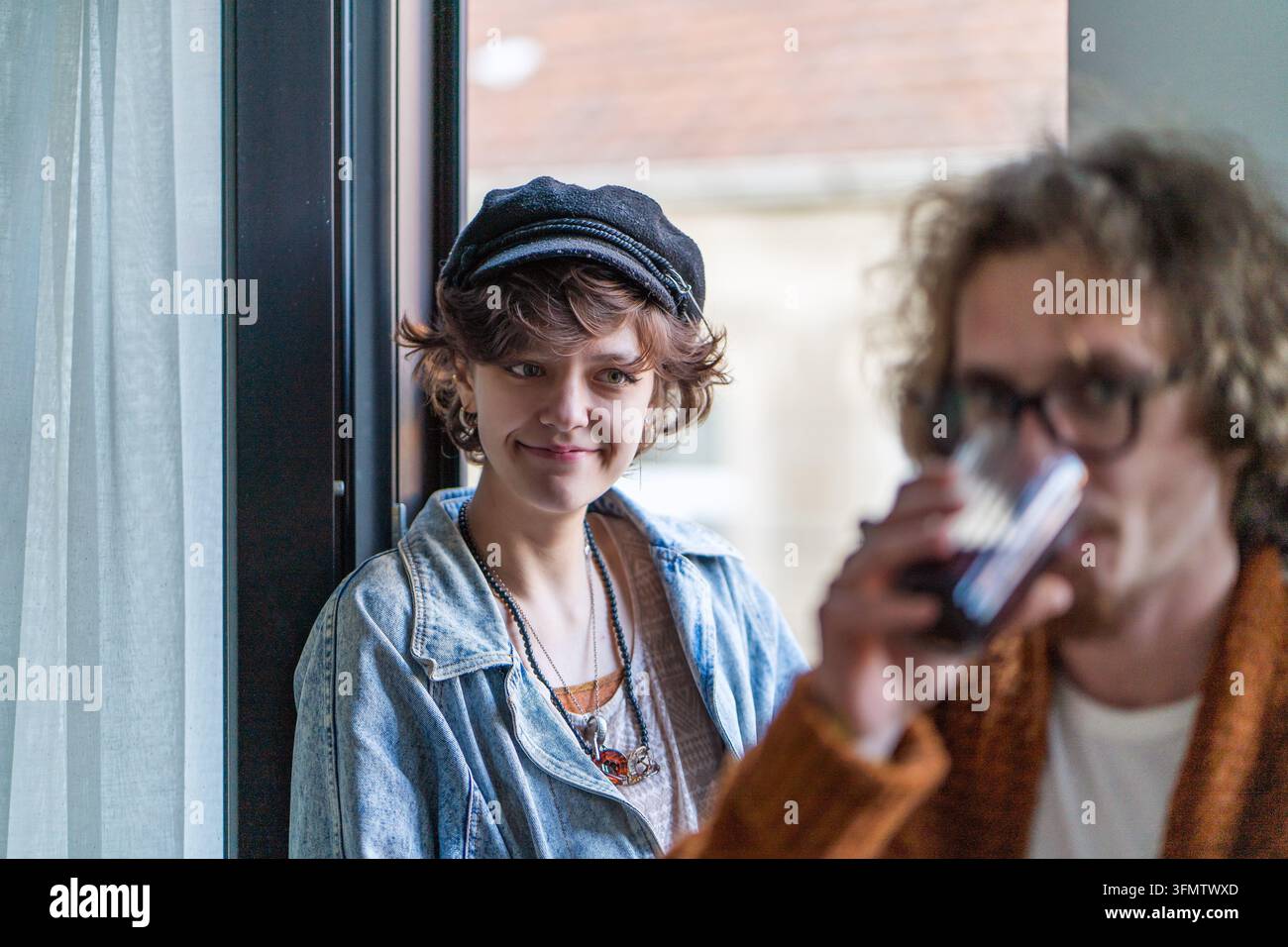 Junges Paar teilt einen entspannten sozialen Moment, während eine lächelnde Frau einen Mann beobachtet, der ein Getränk trinkt, was Partnerschaft, Dating und Verbindung suggeriert. Stockfoto