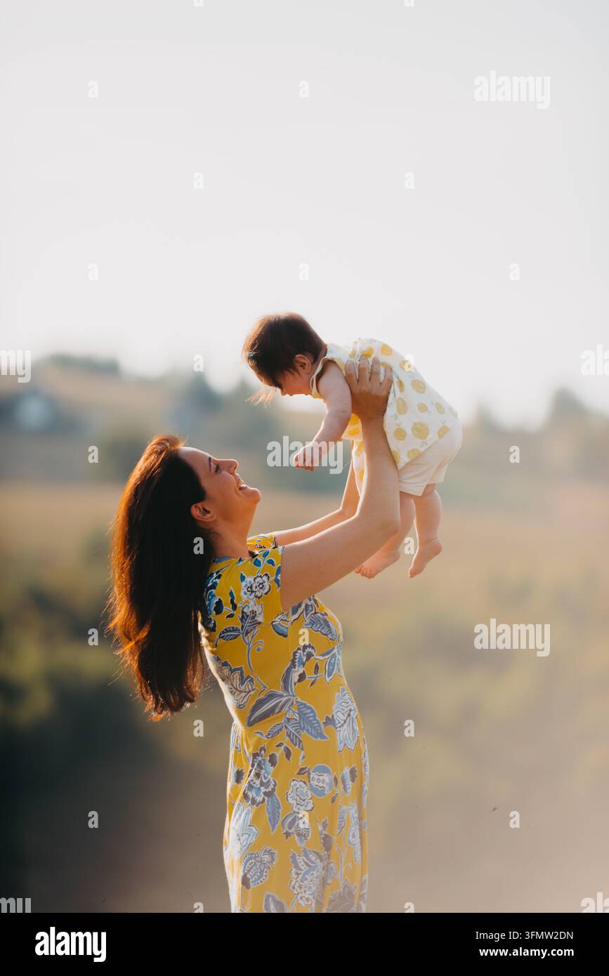 Mutter hebt ihr Baby im Freien in gelbem Kleid während der warmen goldenen Stunde Familienmoment Stockfoto