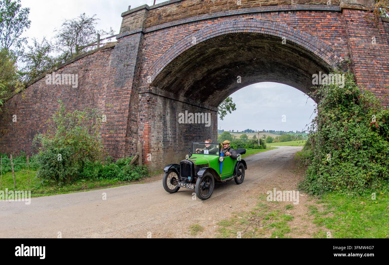 Ein klassisches Cabrio der Austin 7 Chummy fährt eine ländliche Schotterstraße entlang und passiert eine historische gewölbte Ziegelbrücke. Stockfoto