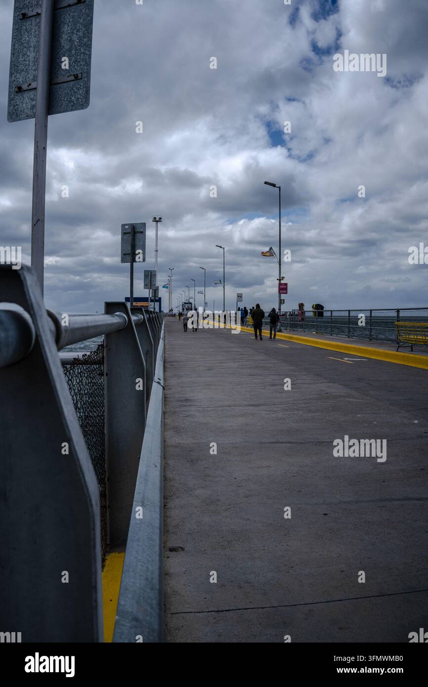 Blick auf den Muelle de Puerto Madryn mit Menschen, die unter einem bewölkten Himmel entlang des Betonstegs in Puerto Madryn, Provinz Chubut, Argentinien spazieren. Stockfoto