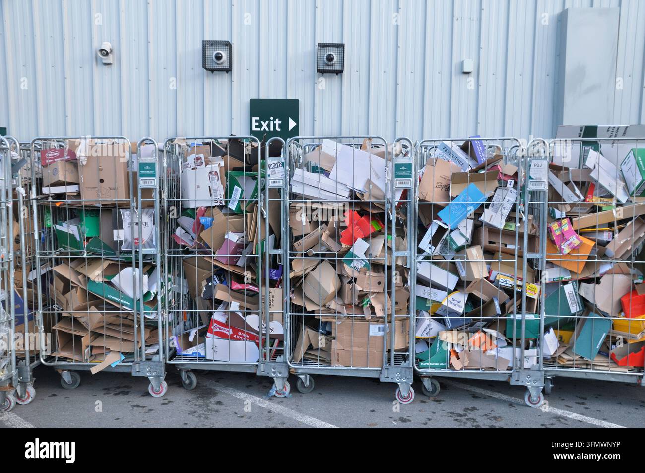 Metallrollenkäfige / 4-seitige Käfige mit Karton- und Verpackungsmüll, die außerhalb eines Supermarktgebäudes unter einem Ausgangsschild aufgereiht sind. Kommerzielles Recycling Stockfoto