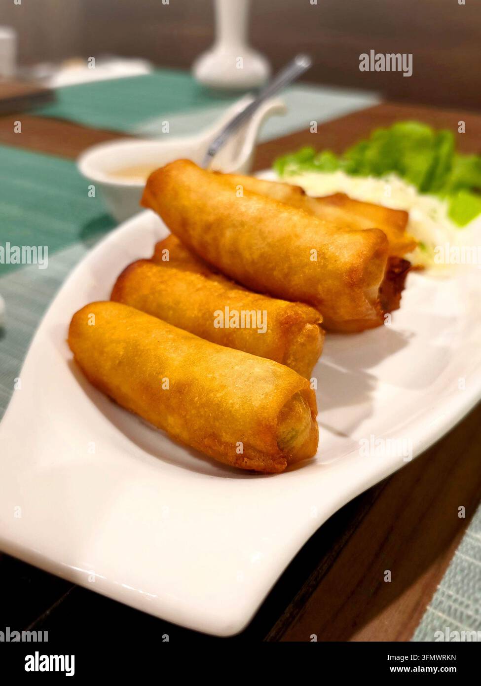Knusprig goldene gebratene Frühlingsrollen serviert auf einem weißen Teller mit süßer Dipsauce und frischem Gemüse in einer Restaurantatmosphäre. - Smartphone-aufgenommenes Stockfoto