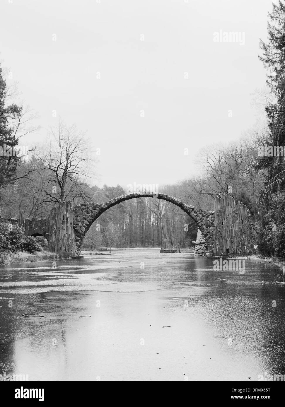 Feinkunst-Monochrom-Fotografie der historischen Rakotz-Brücke, die eine perfekte Kreisreflexion im ruhigen Wasser bildet, umgeben von nebelwaldigem Wald. Stockfoto