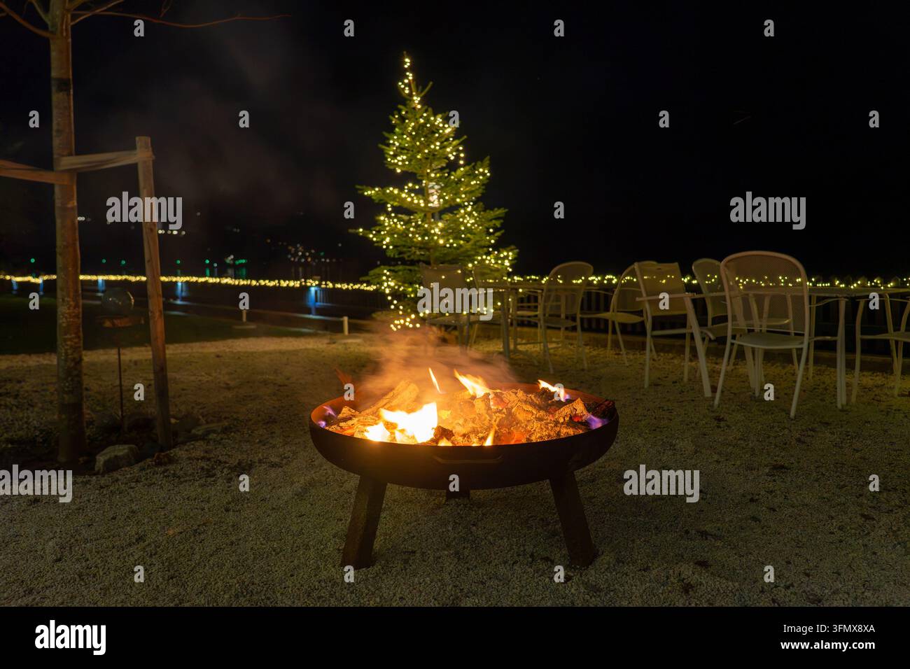 Gemütlicher Feuerkorb und beleuchteter Weihnachtsbaum an einem Alpensee Stockfoto