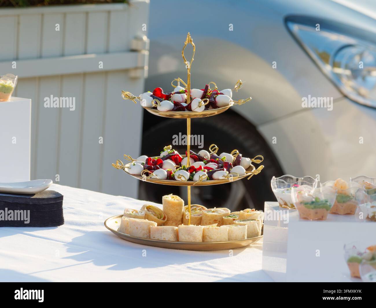 Nahaufnahme eines dreistöckigen goldenen Serviertabletts mit verschiedenen Vorspeisen auf einem weißen Tisch im Freien. Stockfoto