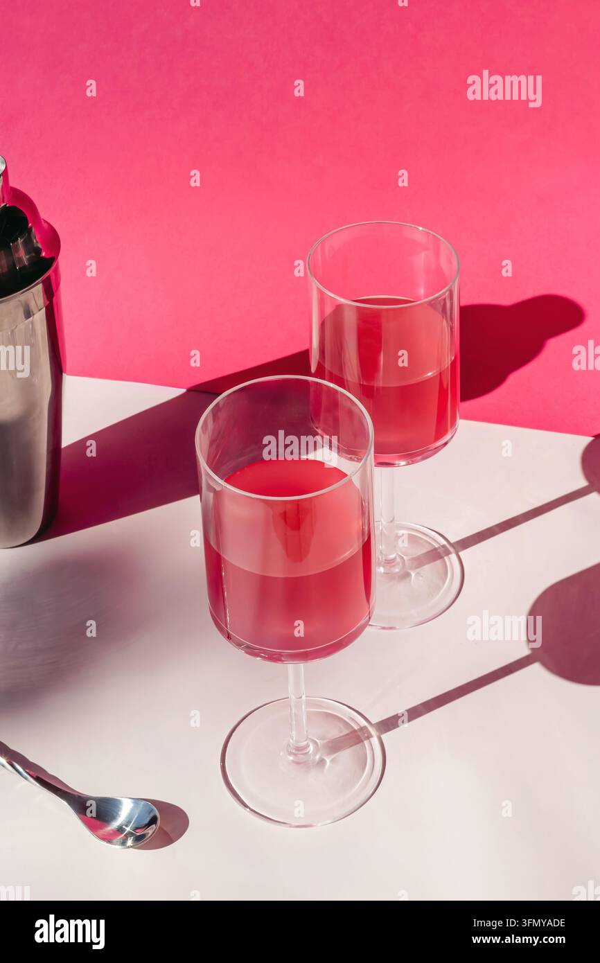 Zwei rosa Getränke in Stielgläsern neben einem Stahl-Cocktailshaker und Barlöffel, helle rosa Wand und grafische Sonnenlichtschatten. Stockfoto