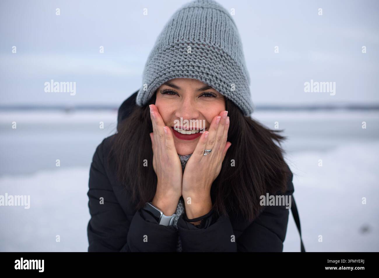Nahaufnahmeporträt einer fröhlichen jungen Frau, die an einem kalten Wintertag am Sylvan Lake, Alberta, Kanada, in die Kamera lächelt und ihr Gesicht hält, um den Ring zu zeigen Stockfoto